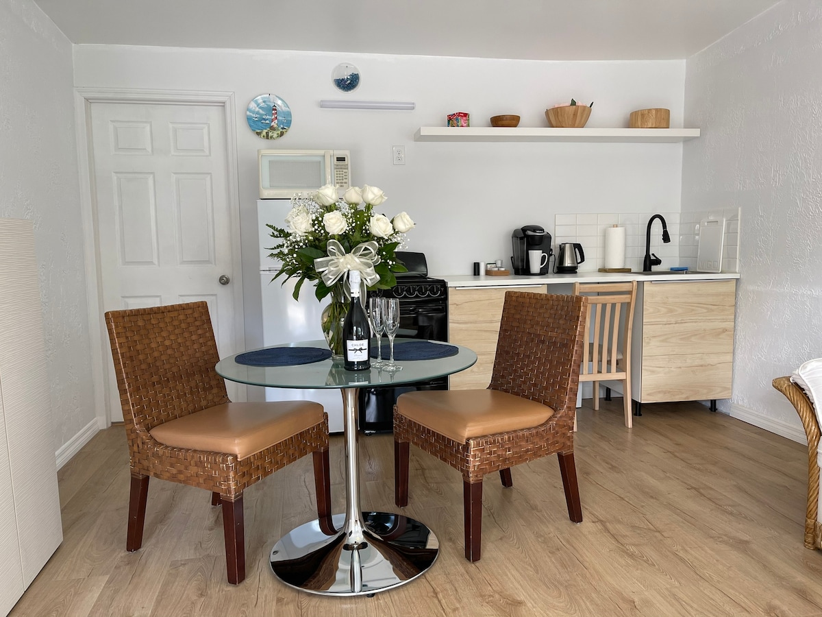 A bright dining area is equipped with a round glass table and two woven chairs. Fresh flowers are arranged in a vase at the center of the table. A modern kitchenette is visible in the background, featuring light cabinetry, a coffee maker, and essential appliances.