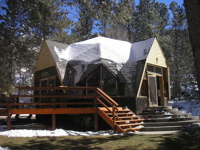 The cabin features a unique architectural design with a sloped roof, surrounded by snow-covered trees. A wooden deck with steps provides access to the entrance, while large windows offer views of the forested area, highlighting the natural setting.