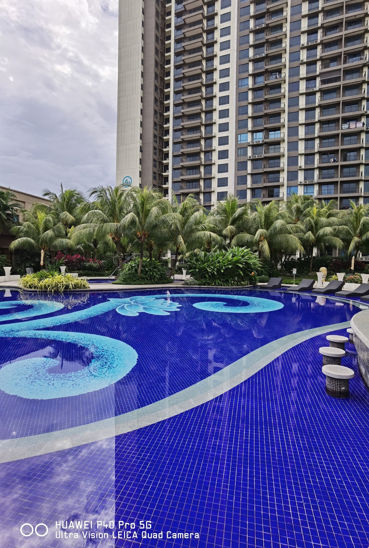 A vibrant swimming pool is surrounded by lush palm trees, with intricate tile patterns on the water's surface. Relaxing seating areas are positioned around the pool, inviting guests to enjoy the tropical surroundings.