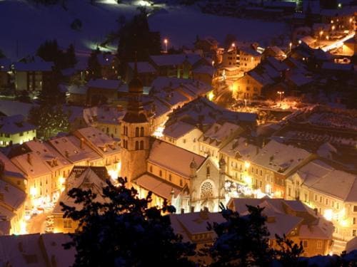 A snowy evening scene of a charming mountain town is visible, with softly lit buildings reflecting warmth against the cold. The historic church, prominent in the center, is surrounded by quaint houses, all covered in a blanket of snow. The tranquil atmosphere is enhanced by twinkling lights throughout the village.
