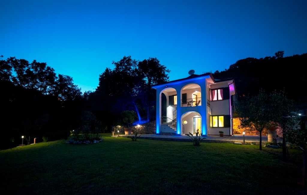The exterior of a two-story house is illuminated at dusk, highlighting elegant architectural features and vibrant lighting. A well-maintained garden extends in front, complemented by trees and shrubs, creating a serene evening atmosphere in a peaceful residential area.