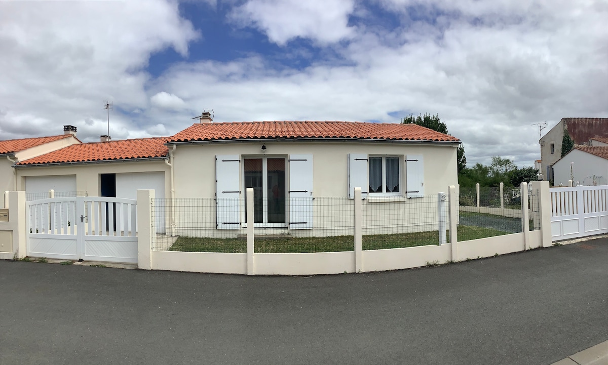 A single-story house is depicted with a red-tiled roof and white shutters. A fenced yard surrounds the property, while a driveway leads to the front entrance. Large glass doors allow natural light to enter, and the sky features a mix of clouds and blue.