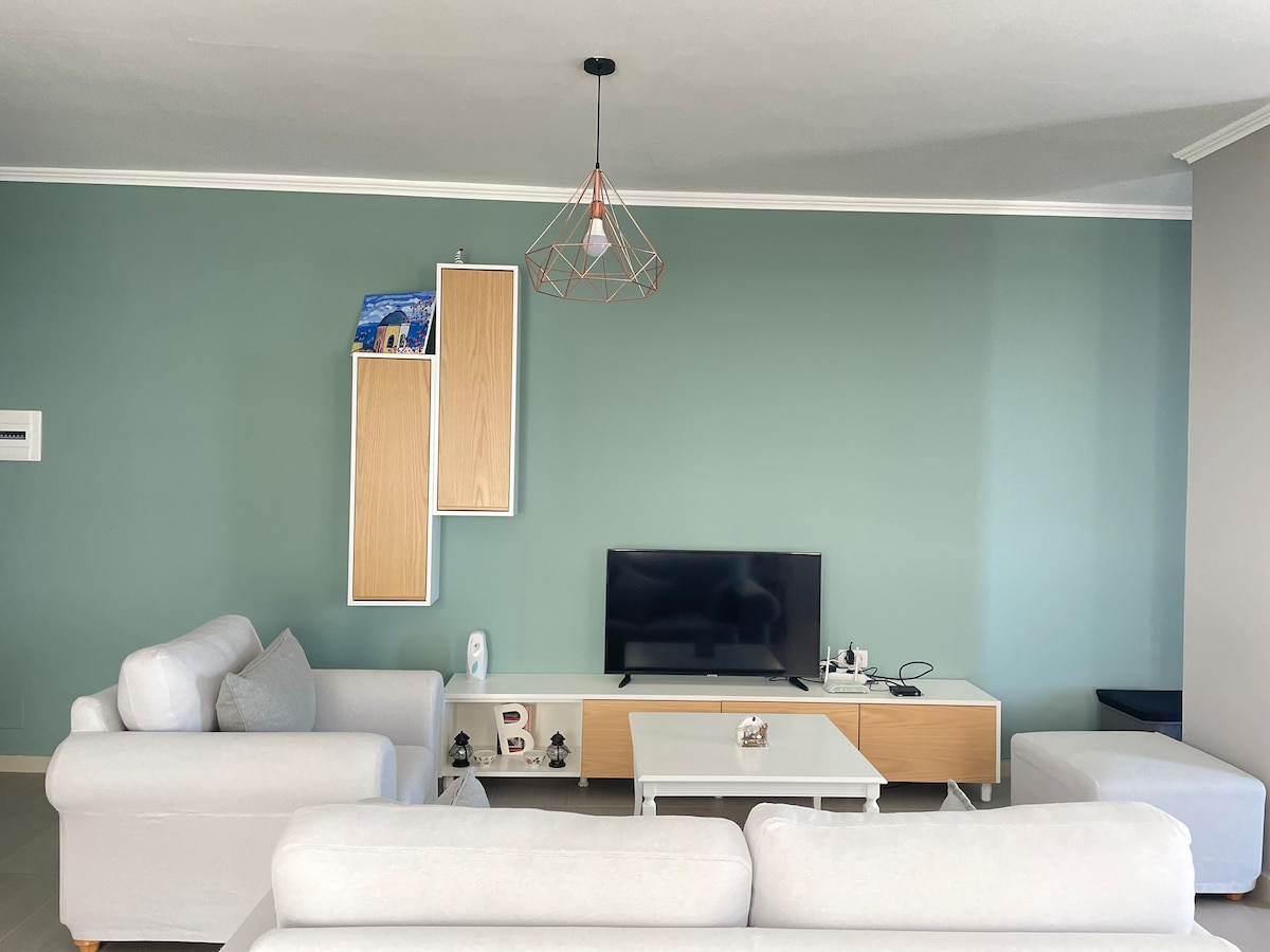 The spacious living area features light-colored sofas arranged around a central coffee table. A large flat-screen TV is mounted against the wall, complemented by modern storage units. A gentle green accent wall offers a calming backdrop, while soft lighting enhances the inviting environment.