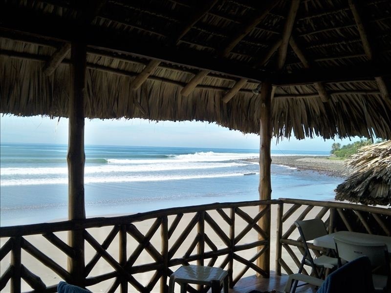 A covered terrace features a thatched roof and wooden railings, providing an unobstructed view of rolling waves and sandy beach. The soothing sound of the ocean is accompanied by a gentle breeze, enhancing the serene environment of this coastal retreat.