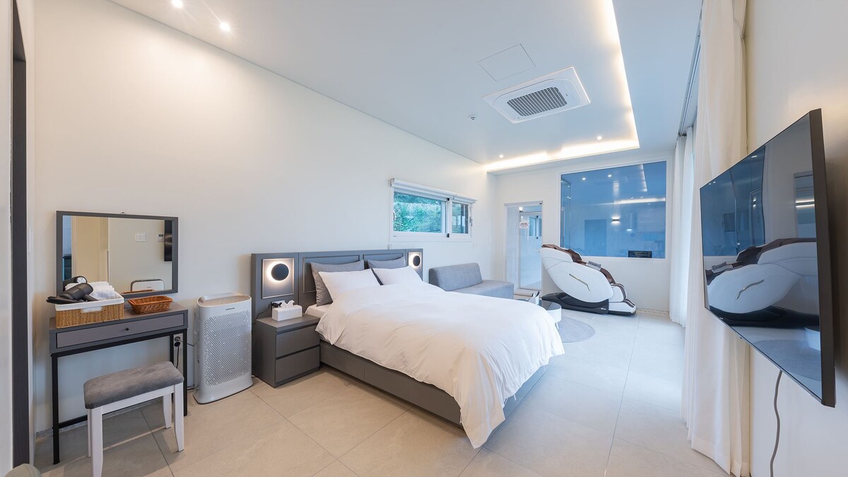 A modern bedroom features a spacious bed with crisp white linens. A wide window allows natural light to illuminate the room. Minimalist decor is complemented by a comfortable seating area and sleek furniture, including a dresser and a flat-screen television mounted on the wall.