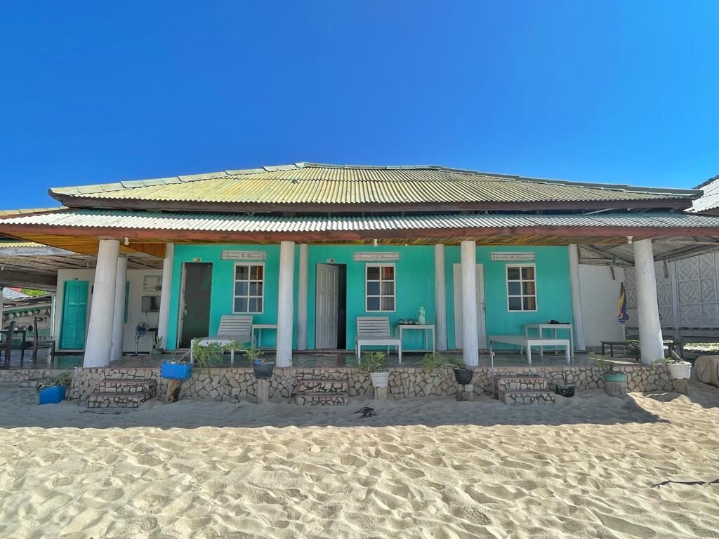 A beachside house features a vibrant turquoise exterior, complemented by white columns. The sandy area in front is lined with various pots and planters. Large windows invite sunlight, while a welcoming porch offers seating options for relaxation.