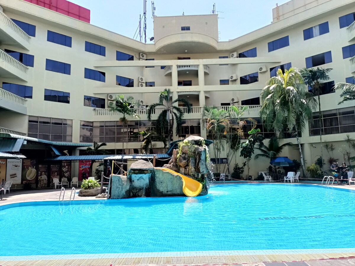 A spacious swimming pool is featured at the center of the resort, surrounded by tropical palm trees. A vibrant water slide and a decorative rock formation are visible, creating a playful atmosphere. Lounge chairs are positioned around the pool area, providing relaxing spots for guests.