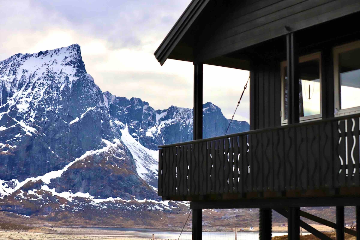 The exterior of the cabin showcases a dark wooden facade with a patterned railing. Snow-capped mountains rise majestically in the background, providing a stunning contrast to the cabin. A tranquil landscape is visible, enhancing the cabin's inviting presence in this natural setting.
