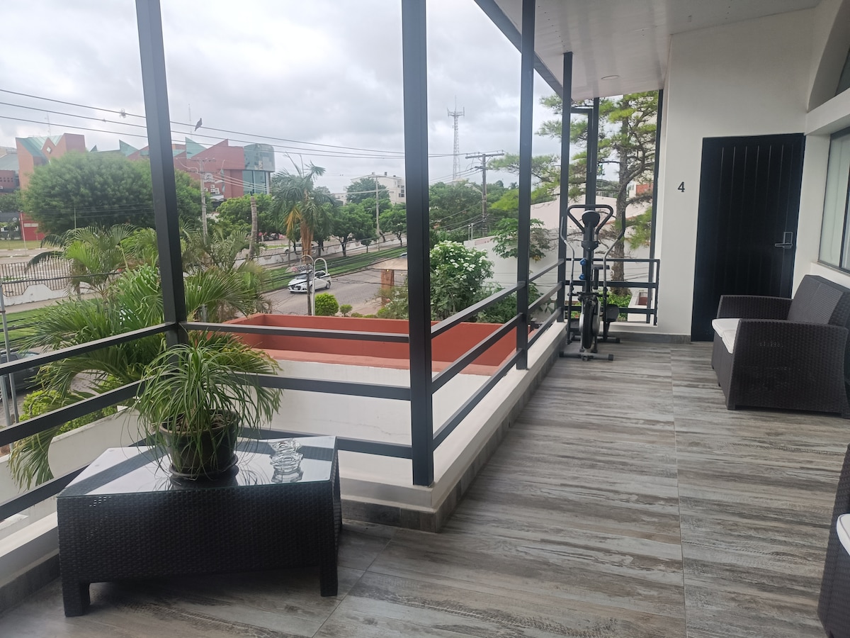 A spacious balcony features modern furnishings, including a sleek table and two lounge chairs. Potted plants add greenery, and a fitness machine is positioned nearby. The view includes tree-lined streets and nearby buildings, enhancing the open and airy feeling of the space.