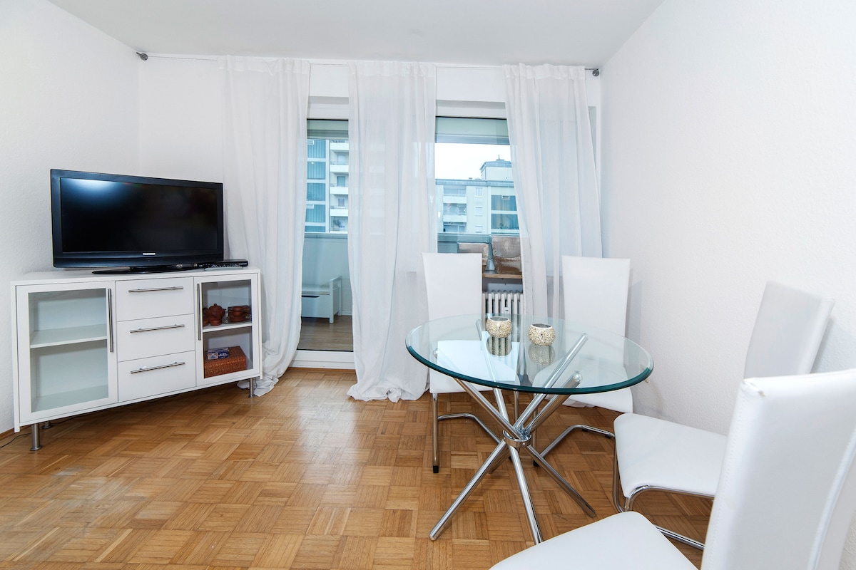 A bright dining area features a circular glass table surrounded by four white chairs. A television is positioned on a sleek cabinet, with sheer white curtains framing a window that allows natural light to fill the space.