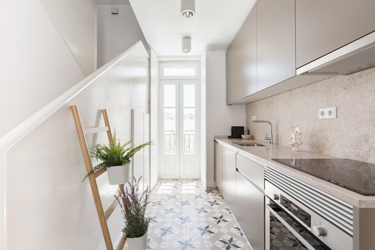 A modern kitchen is presented with sleek cabinetry and stainless-steel appliances. Natural light floods through large windows, highlighting the intricate tile flooring. A small ladder shelf stands against the wall, adorned with potted plants for a touch of greenery.