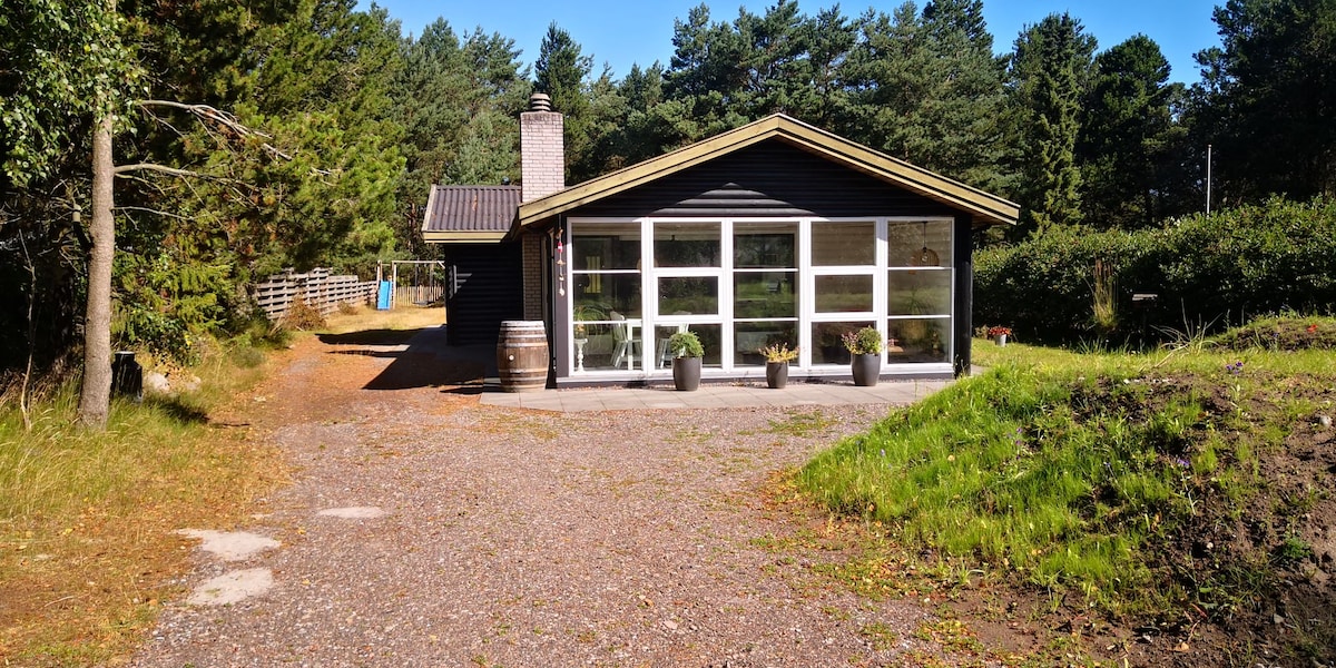A single-story summer house is set amid lush greenery, featuring large windows that offer ample natural light. The exterior is painted in dark tones, complemented by several outdoor planters. A gravel pathway leads to the entrance, which includes rustic details like a barrel.