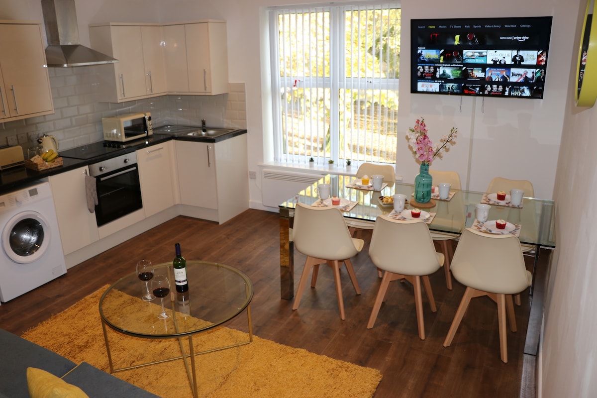 A modern living area features a glass dining table with white chairs, elegantly set with dishes and drinks. A smart TV is mounted on the wall, showcasing various entertainment options. Sunlight filters through large windows, illuminating the stylish kitchen and cozy seating area.