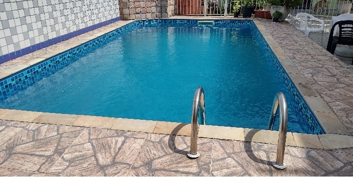 A clear swimming pool is bordered by light-colored tiles, with silver handrails positioned at one end. The pool's surface reflects the sky, while a stone wall and lush greenery provide a serene backdrop.