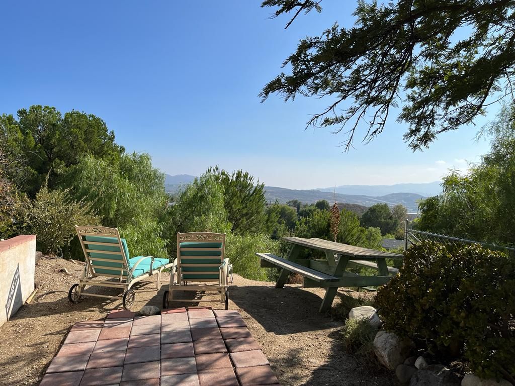 Two lounge chairs are positioned on a stone patio, providing a view of the surrounding hills and greenery. A picnic table is visible nearby, surrounded by lush foliage and a clear sky, creating an inviting outdoor space for relaxation.
