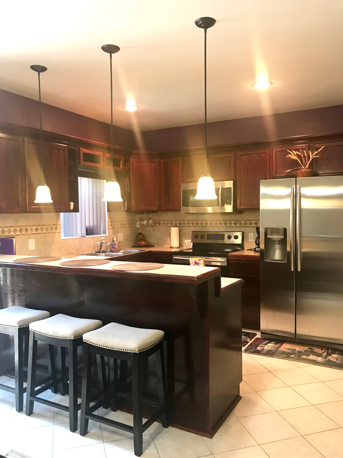 The kitchen features dark wood cabinetry complemented by stone countertops. A central island with three bar stools provides casual seating. Pendant lights illuminate the space, and modern appliances, including a stainless steel refrigerator, are positioned for convenience. Natural light enters through a nearby window.