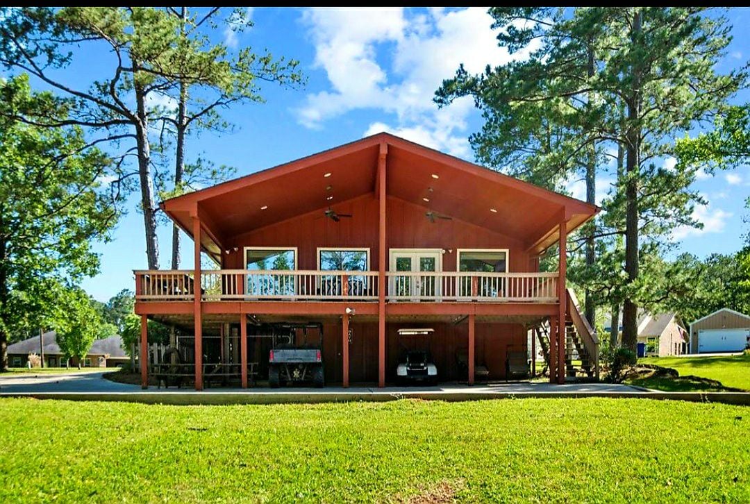 The exterior of Water's Edge, a red two-story home, is showcased. Surrounded by a yard and trees, the spacious deck provides various outdoor seating options, and large windows offer views from the upper level. The area in front accommodates parking for vehicles and watercraft.