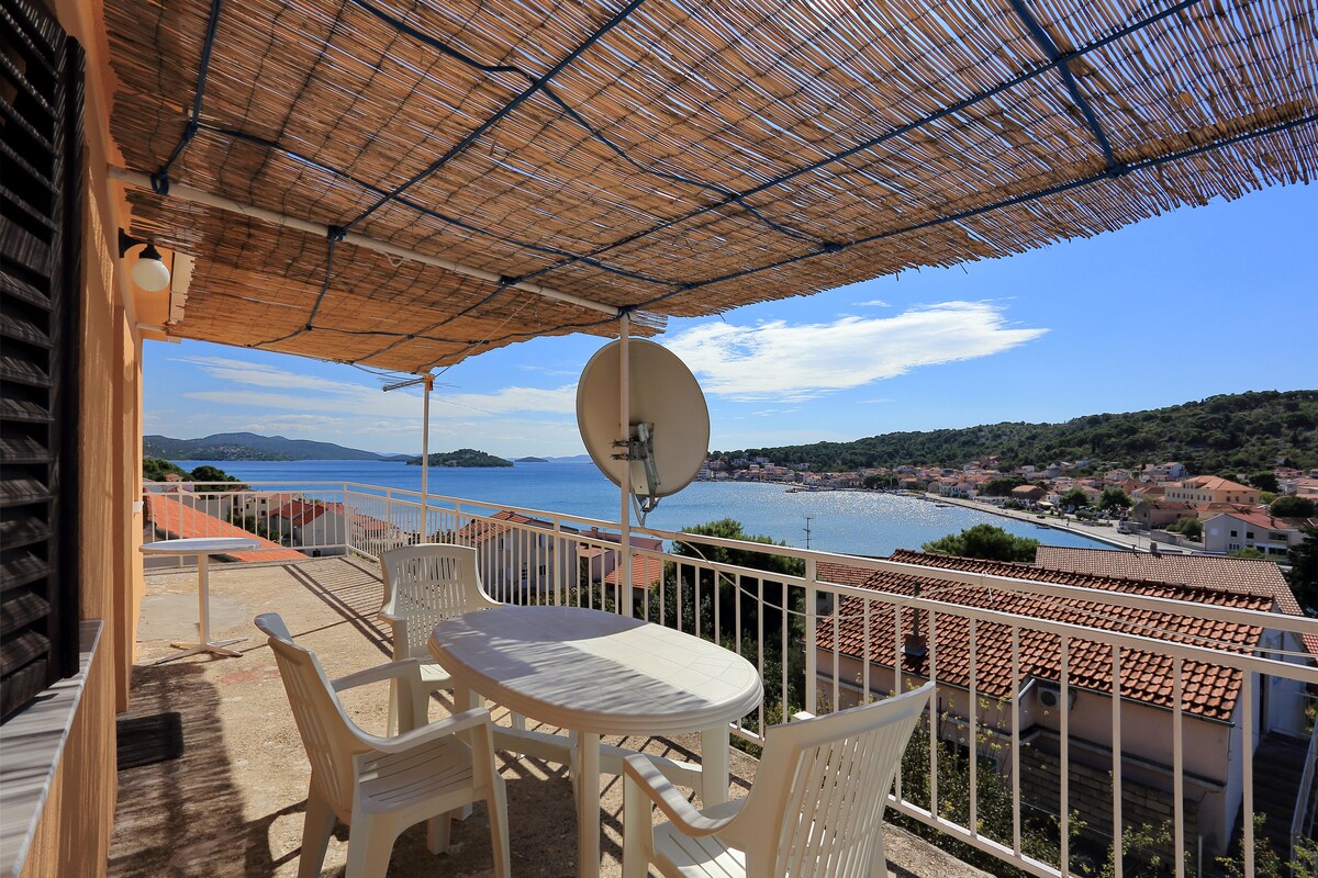 A spacious terrace is covered by a reed awning, offering a view of the village and surrounding sea. A round table with four white chairs is positioned on the terrace, creating an inviting outdoor space for relaxation.