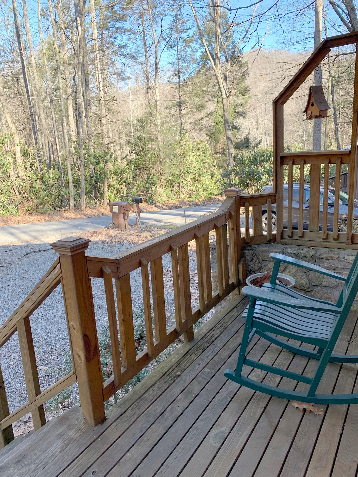 A back porch is featured with wooden railings and flooring, overlooking a serene wooded area. A rocking chair is positioned for relaxation, and a gravel driveway is visible nearby. The surrounding trees provide a natural backdrop, enhancing the tranquil outdoor space.