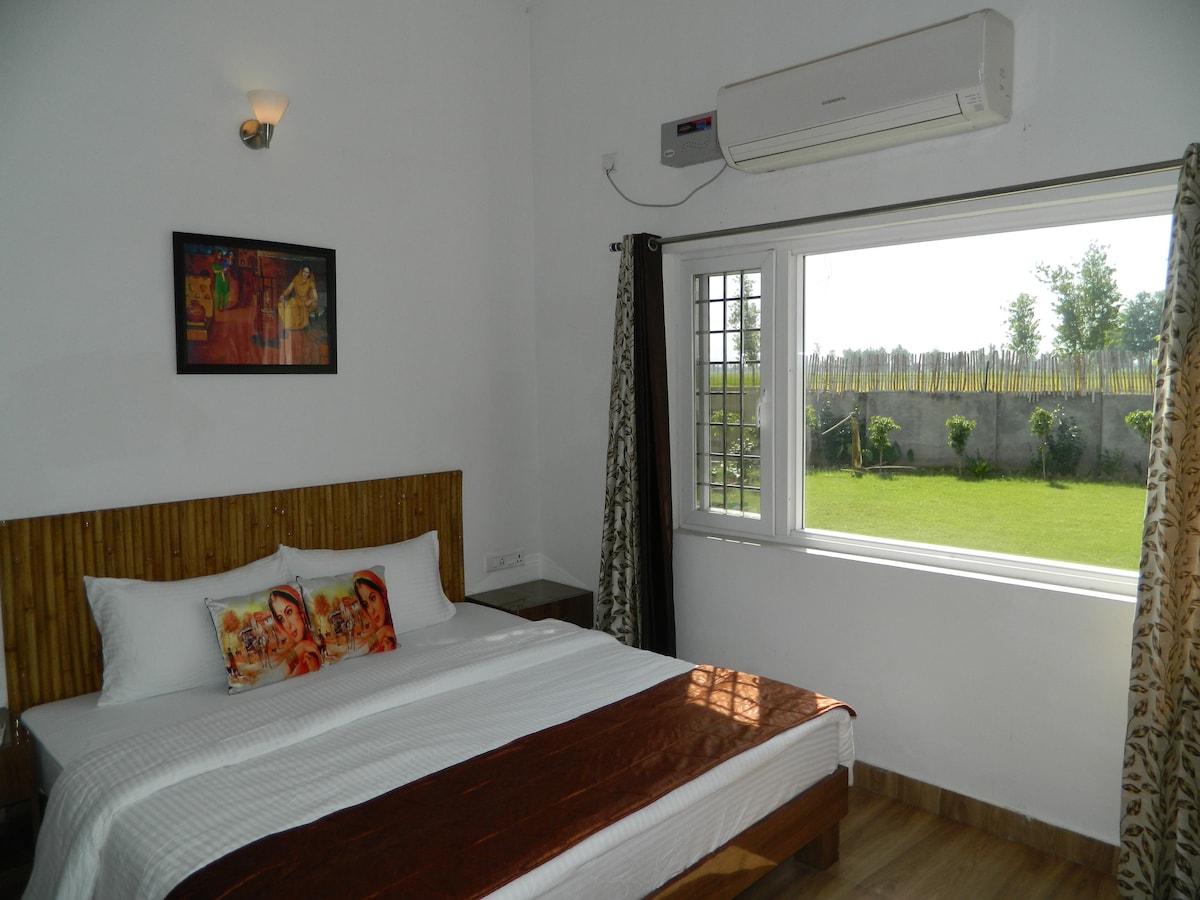 A well-furnished room features a spacious bed with decorative pillows and crisp linens. A large window offers a view of the green outdoors, brightening the space. The room is equipped with air conditioning, and artistic wall decor adds a touch of style.