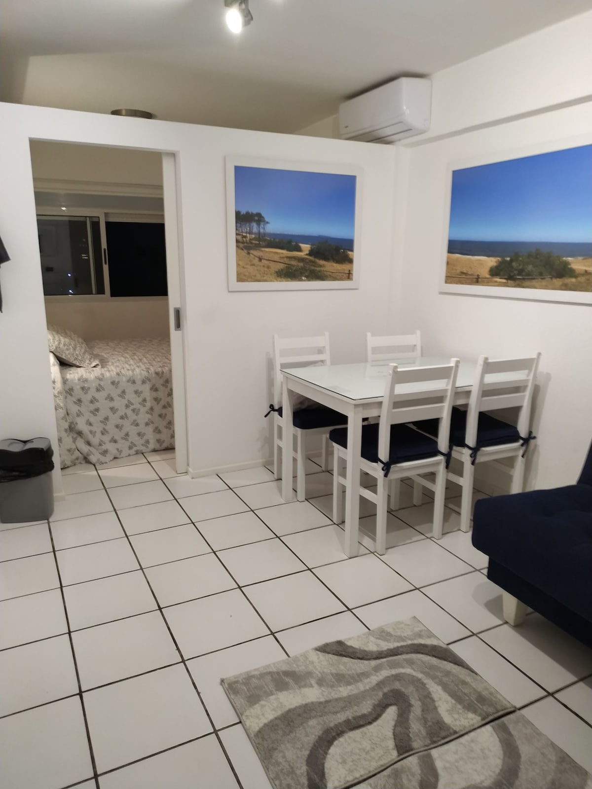 A bright living area features a white dining table surrounded by four chairs. Soft blue tones are introduced with a plush sofa. Two large windows reveal views of a coastal landscape. A cozy sleeping space is visible through a partially open door.