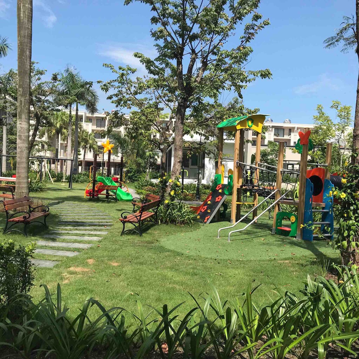 A children's playground is visible in a spacious outdoor area, featuring a colorful play structure with slides and climbing elements. Benches are arranged around lush green grass and trees, providing shaded seating for adults. Clear blue skies offer a bright and inviting atmosphere.