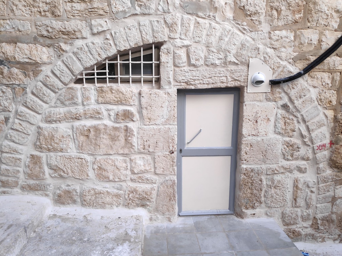 A traditional stone wall is featured, characterized by large, rough-hewn stone blocks. An arched doorway is visible, with a simple gray door and a security grate positioned above. The ground is paved with smooth stone tiles, emphasizing the rustic charm of the architecture.
