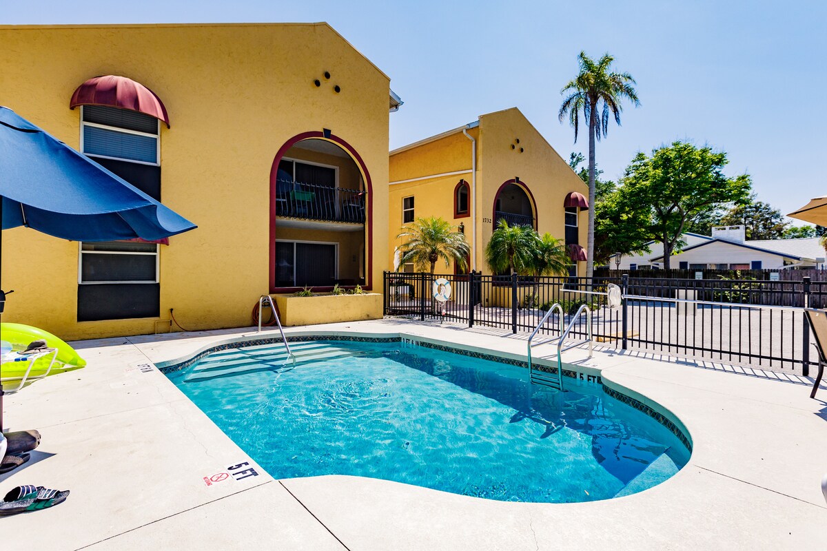 A shared swimming pool is surrounded by a fenced area, with sun loungers and umbrellas nearby. The vibrant yellow building creates a cheerful backdrop, while palm trees provide a tropical touch. Clear skies enhance the overall bright and inviting atmosphere of the space.