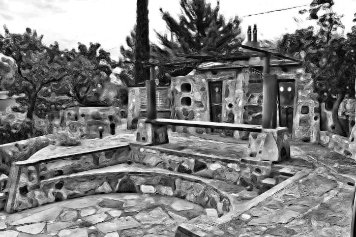 An outdoor area is surrounded by natural stone walls and pathways, featuring a tiered seating arrangement made of stone. A shaded pergola overhead provides cover, while trees and shrubs are present throughout the space, contributing to a rustic and earthy environment.