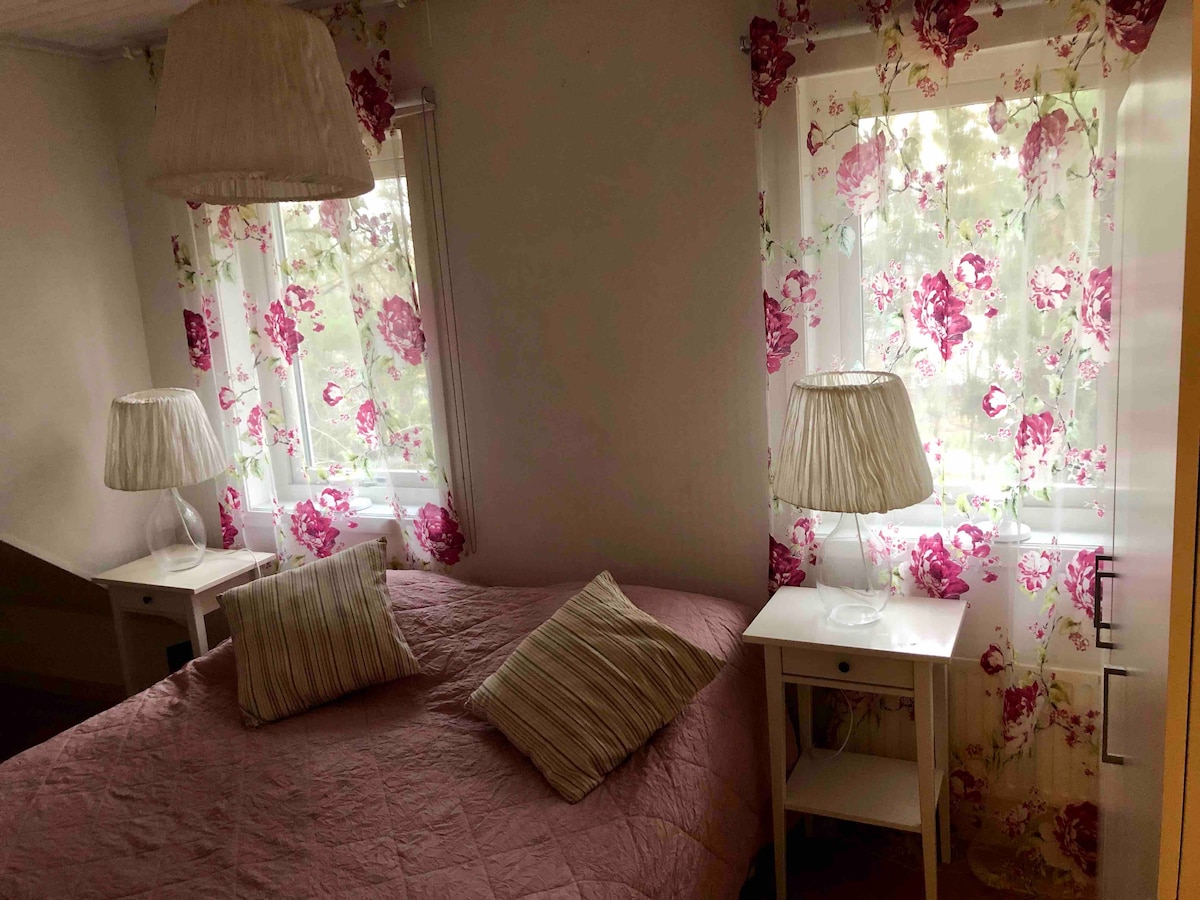 A cozy room features a spacious bed covered in a light pink bedspread, flanked by two white nightstands. Soft floral curtains adorn the windows, allowing dappled light to enter. Two lamps with pleated shades offer additional illumination, creating a calming ambiance.