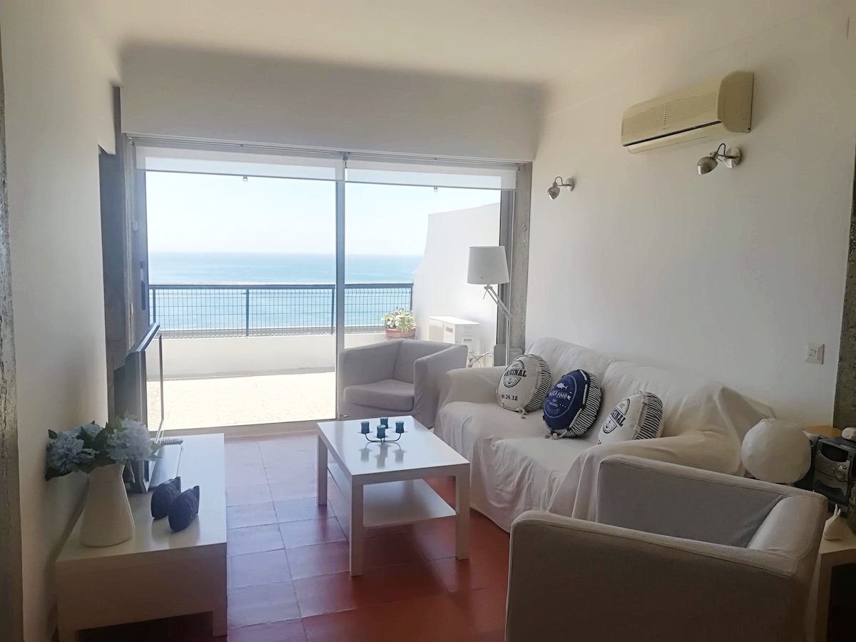 A bright living area features comfortable seating arranged around a white coffee table, with decorative objects positioned on it. Large glass doors provide a panoramic view of the ocean and lead to an outdoor terrace. Soft natural light fills the room.