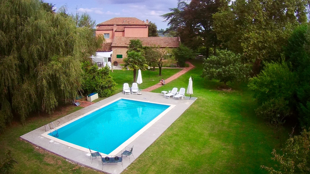 An aerial view reveals a rectangular swimming pool surrounded by manicured green lawns and shaded by trees. Several sun loungers and chairs are positioned nearby, with pathways leading through the lush garden. The main house is visible in the background, framed by foliage.
