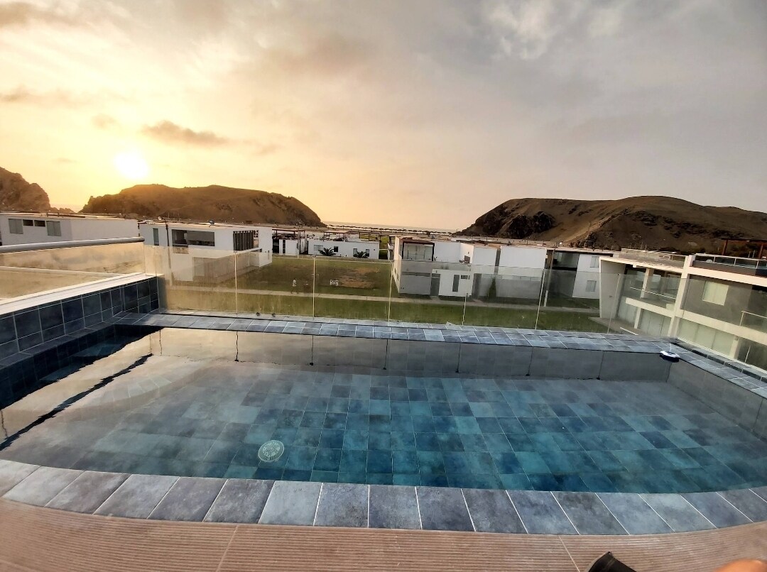 A large swimming pool is shown, featuring a tiled surface reflecting the sunset. Surrounding glass barriers provide an unobstructed view of the landscape, including hills and the ocean at a distance, enhancing the serene atmosphere.