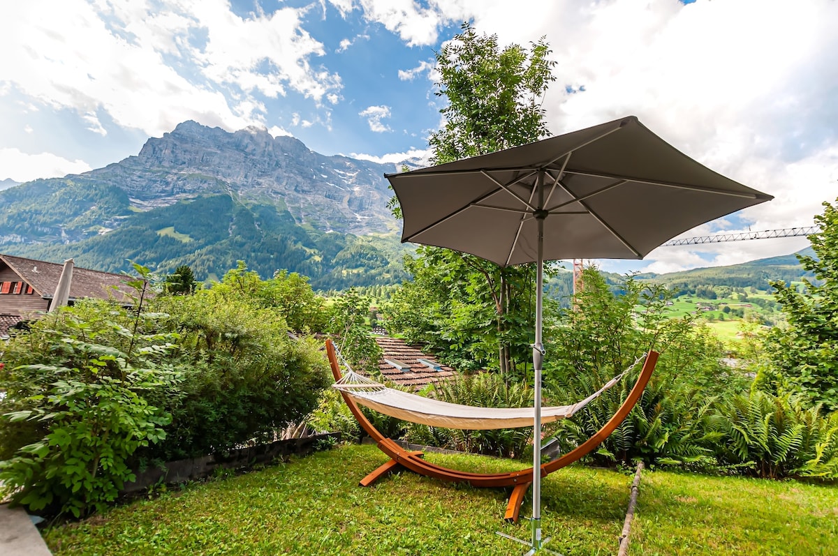 A serene garden area is presented, featuring a wooden hammock supported by curved stands. A large umbrella provides shade, while vibrant greenery surrounds the space. The majestic mountain landscape is visible in the background, enhancing the outdoor setting.