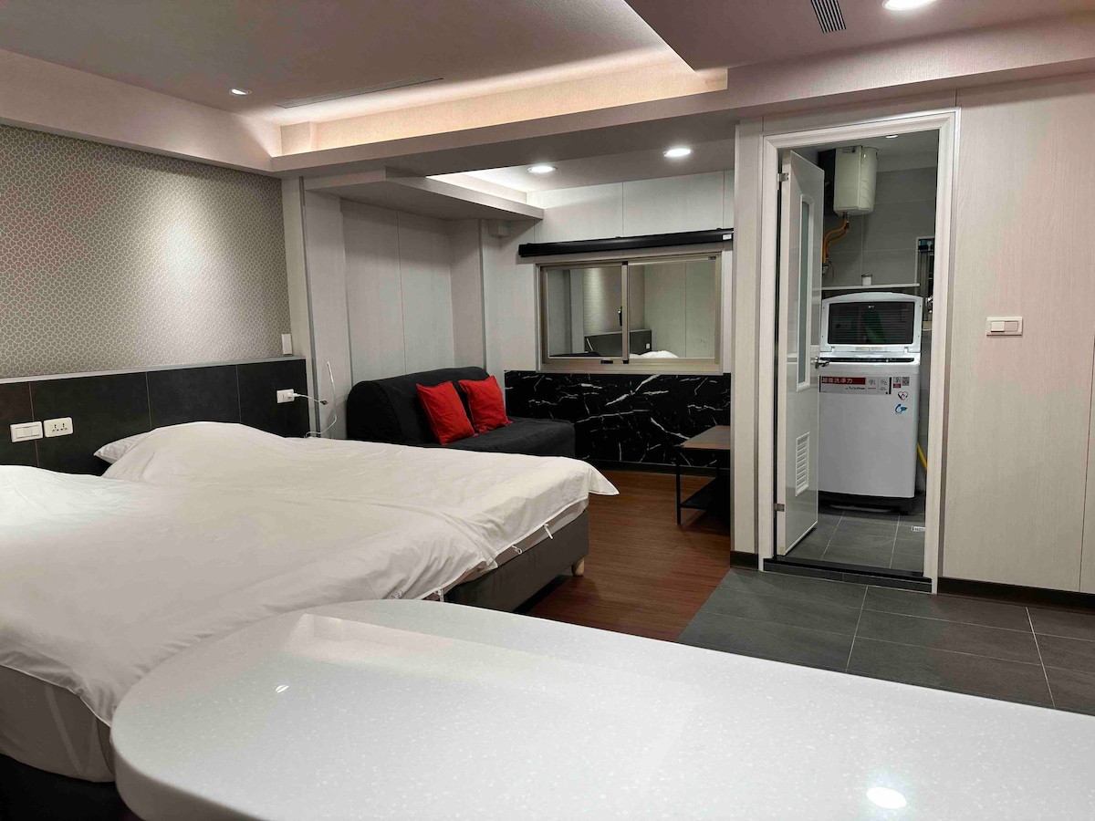A spacious room is presented, featuring two single beds that can be combined. A sofa with red cushions offers additional seating. A door leads to a laundry area, and a sleek countertop is visible in the foreground. The overall lighting is bright and inviting.
