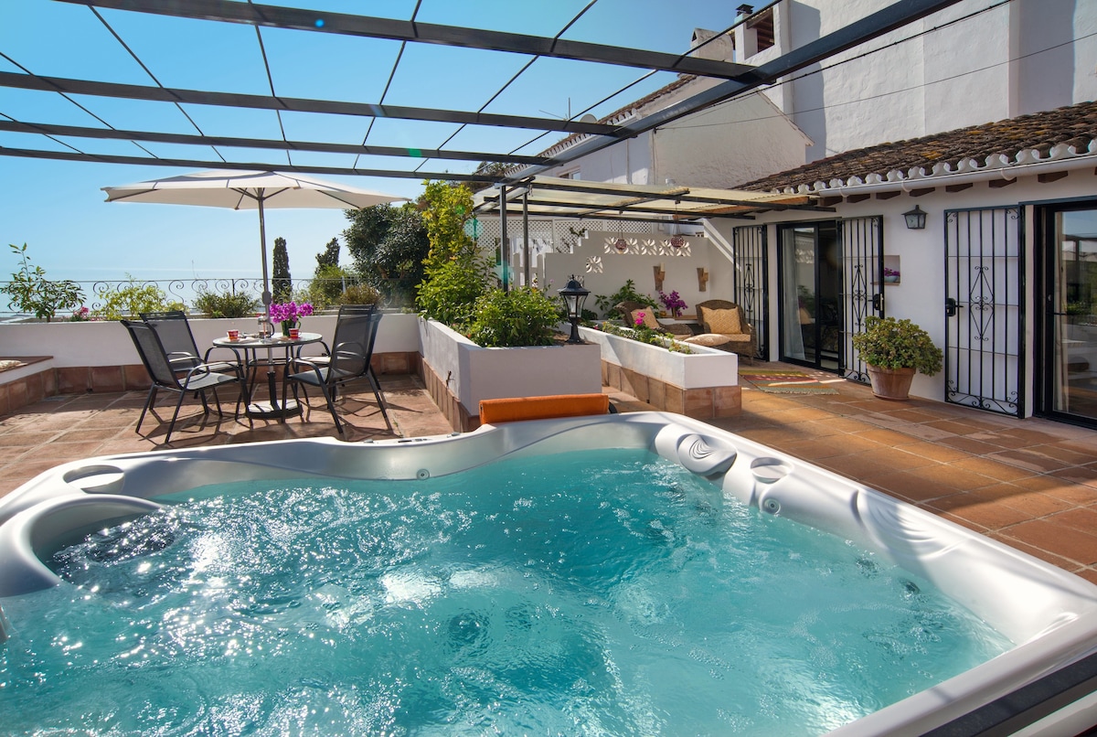 A private outdoor patio area features a hot tub surrounded by lush greenery. A shaded dining table with chairs invites alfresco meals, while a sun umbrella provides additional comfort. The scene is complemented by views of the sky and surrounding landscape.