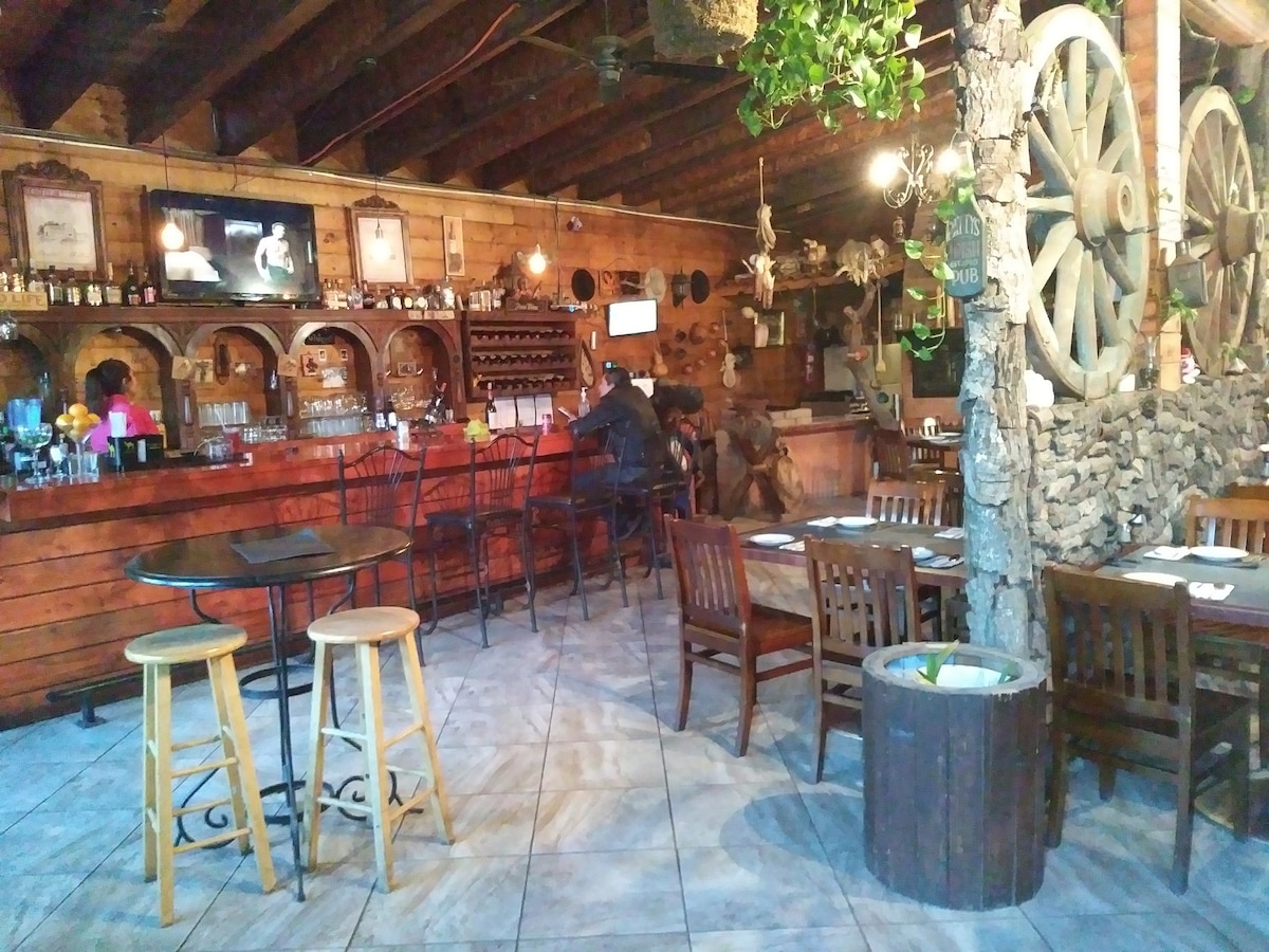A rustic dining area is showcased with wooden beams overhead and stone accents. Tables and chairs are arranged around the space, with a long bar visible at the back. Decorative elements include trees and wagon wheels, creating a warm, inviting atmosphere for guests.