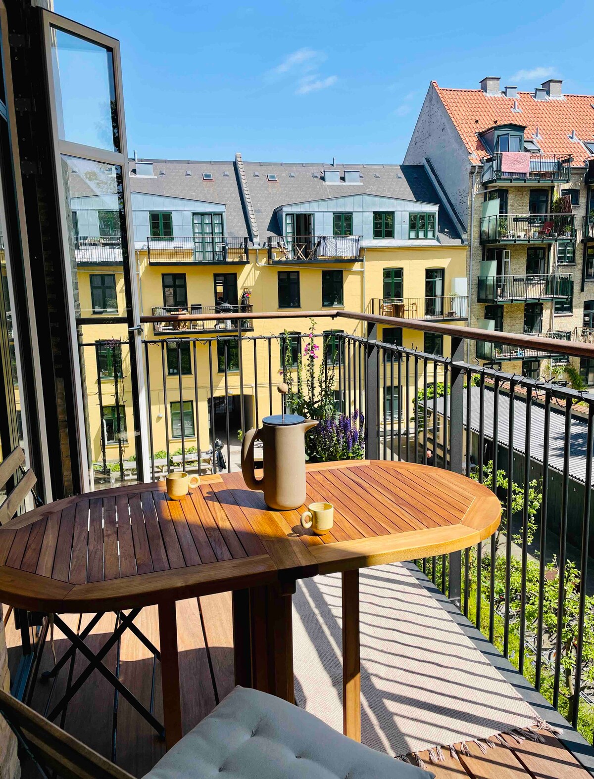 A private balcony is featured, furnished with a wooden table and chairs, complete with a teapot and cups. The view includes colorful buildings and greenery, with sunlight streaming across the space, creating a bright and open atmosphere.