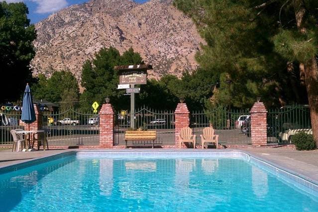 A clear swimming pool reflects the surrounding landscape, framed by a fence and brick columns. Sun loungers are positioned nearby, offering a place to relax. Lush trees provide shade, while distant mountains form a natural backdrop under a bright blue sky.
