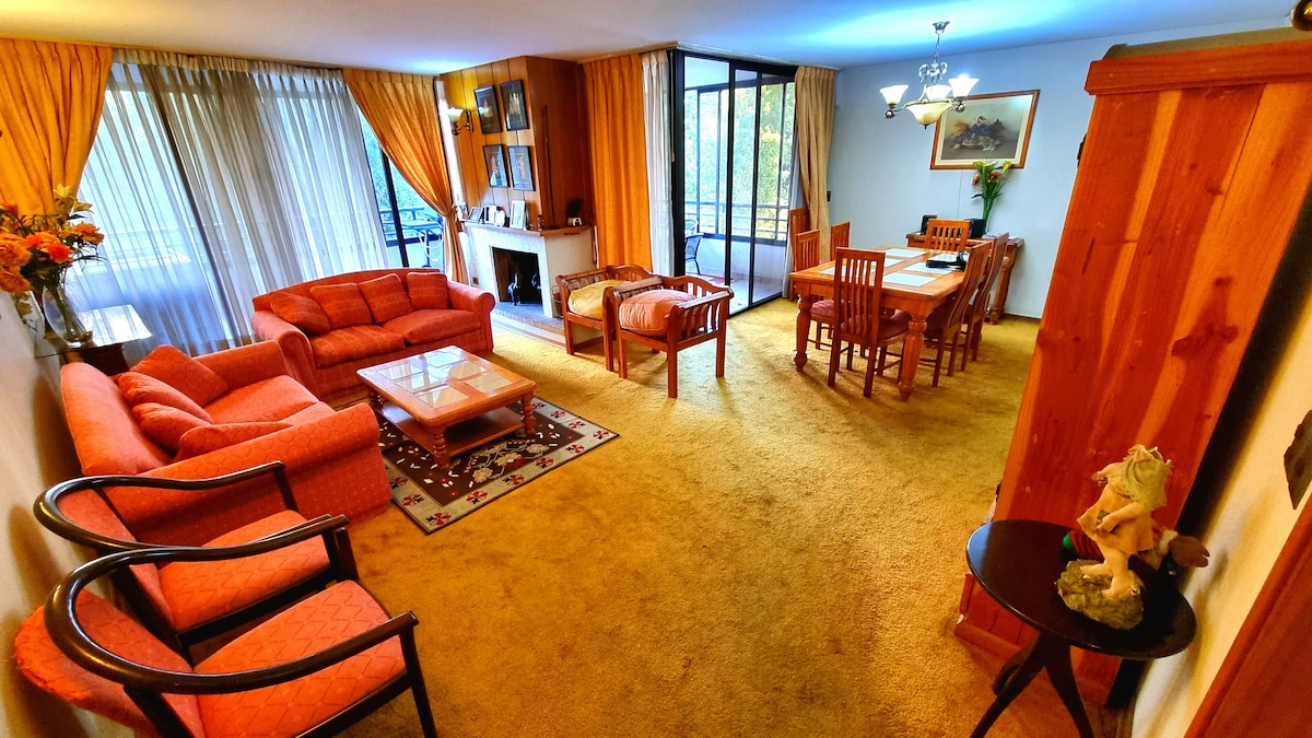 A spacious living area features a combination of soft seating in warm tones, accented by a glass coffee table. Natural light floods through large windows with sheer curtains, illuminating the cozy atmosphere. A dining table is visible in the background, complemented by wooden chairs.