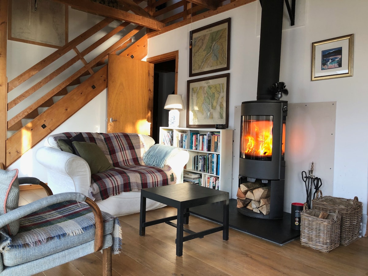 A cozy living area features a wood-burning stove emitting warmth, surrounded by a plaid sofa and a small black coffee table. Nearby, a shelf filled with books adds to the inviting atmosphere, while framed maps and artwork adorn the walls.
