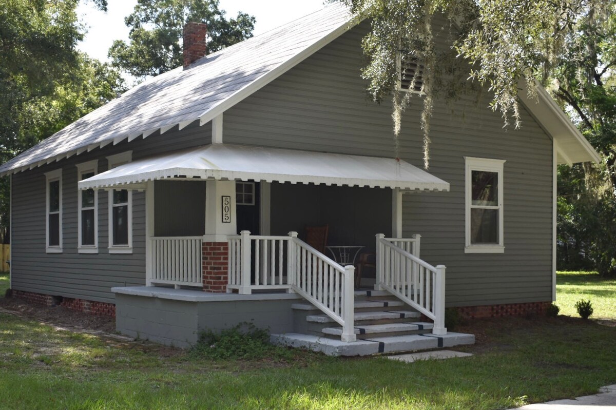 The exterior of a historic bungalow is depicted, featuring a welcoming porch with a slatted awning. Light gray siding complements the brick foundation, while large windows provide views of the surrounding green space shaded by trees.