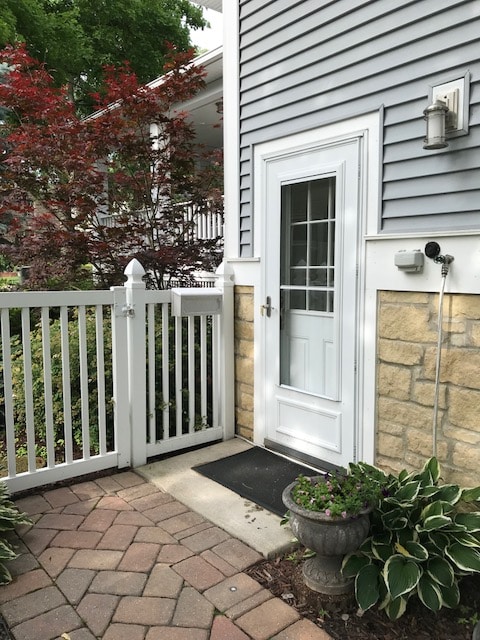 A private entrance is seen, featuring a white door framed by a stone wall and surrounded by green foliage. The patio is paved with brick, and a planter with leafy plants adds a touch of greenery near the door.