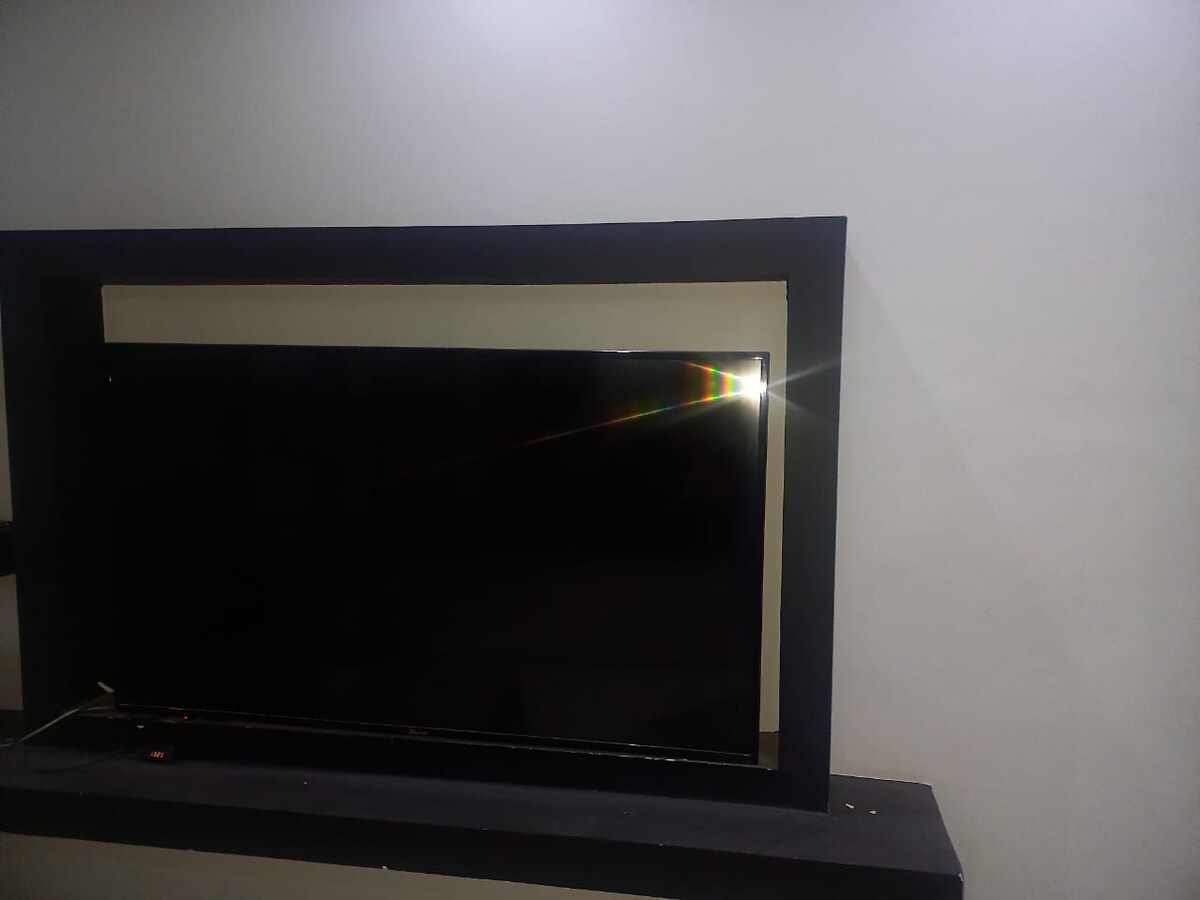 A modern flat-screen television is mounted on a sleek black shelf, set against a smooth, light-colored wall. The television screen reflects a subtle glare, indicating the presence of light in the room.