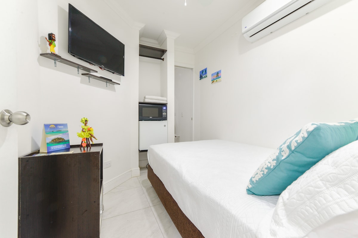 A cozy room features a spacious bed with white bedding, complemented by a decorative blue cushion. A television is mounted on the wall above a small shelf. A kitchenette area includes a microwave and refrigerator. Natural light brightens the room, creating a simple and functional space.