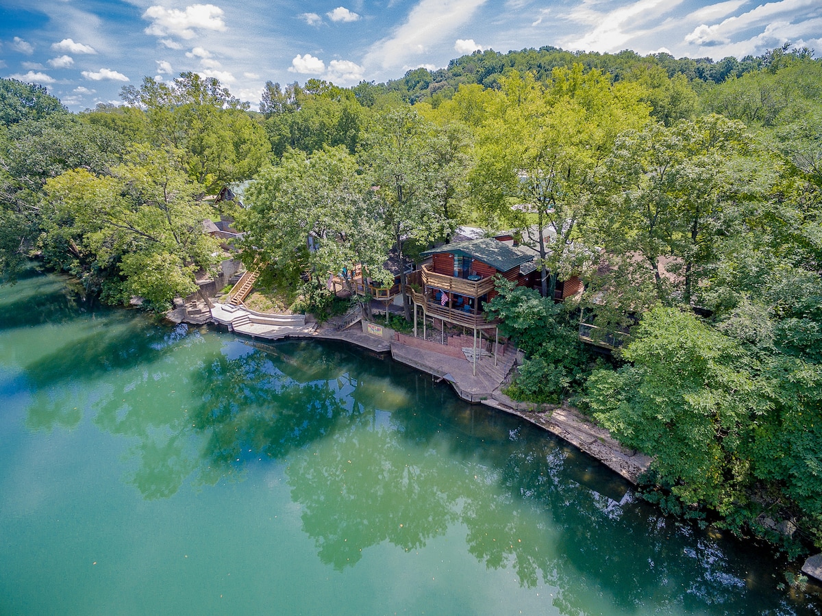 An aerial view of a log cabin situated alongside a serene river, surrounded by lush greenery. The cabin features a spacious deck, providing direct access to the water, with steps leading down to the riverbank. The calm waters reflect the blue sky and tree canopy.