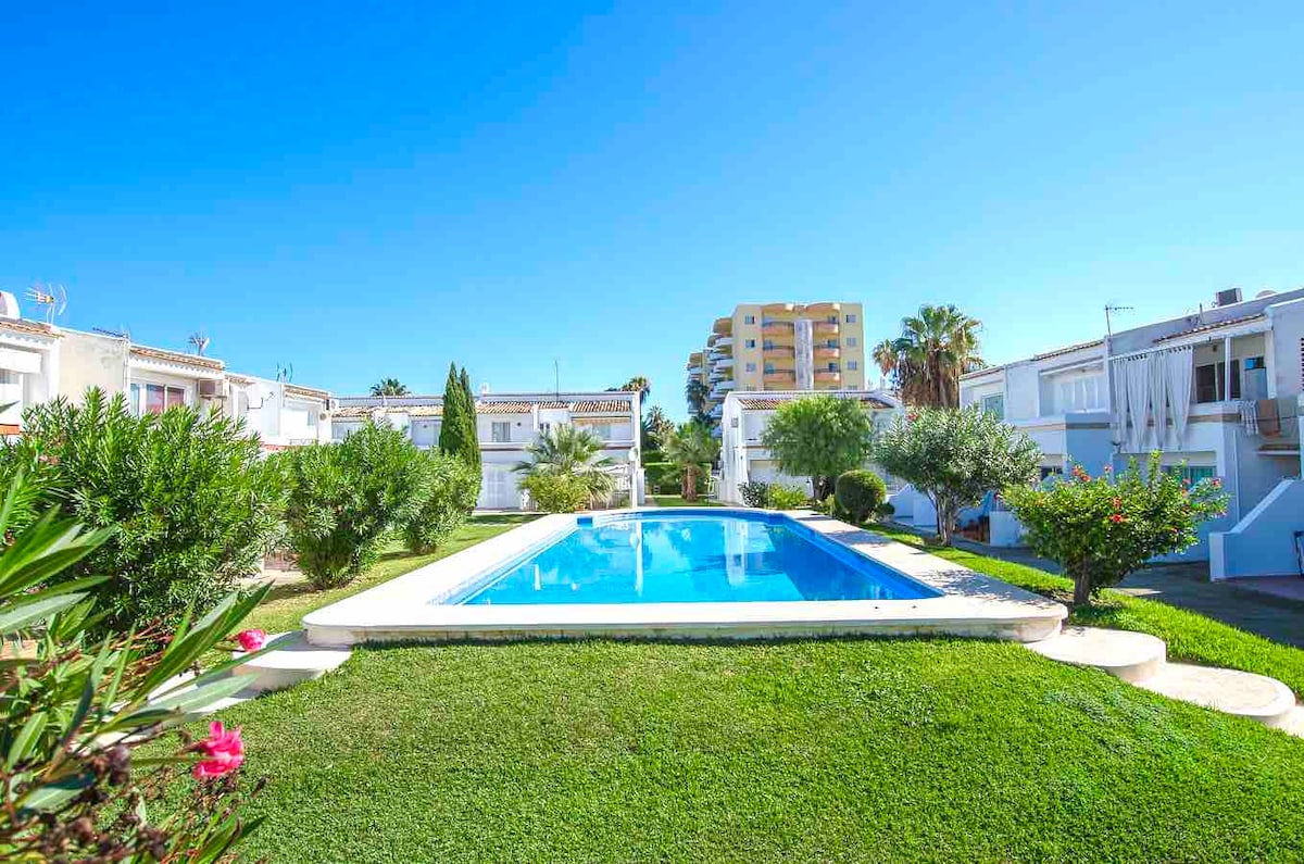 A bright community pool is surrounded by well-maintained lawns and lush greenery. Palm trees are visible in the background, alongside residential buildings. The clear blue sky contributes to the inviting atmosphere of the outdoor space.