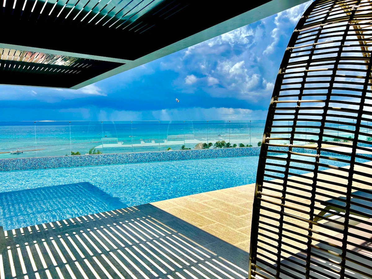 A serene infinity pool appears to extend into the ocean, framed by a modern structure. The tranquil water reflects the sky, while a stylish seating area is positioned under a contemporary awning, providing shade and a view of the Caribbean Sea.