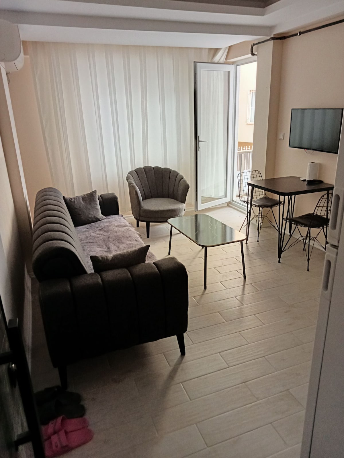 A cozy living area is furnished with a plush gray couch and two stylish chairs, creating a comfortable seating arrangement. A glass coffee table sits in the center, complemented by a small dining table with two chairs nearby. Light enters through a glass door leading to a balcony.