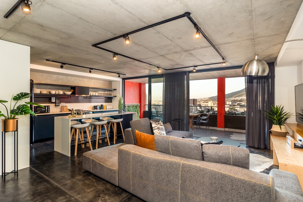 An open-plan living area features a modern sofa facing a large television. A kitchen with bar stools is visible in the background, along with shelves displaying kitchenware. Floor-to-ceiling windows provide natural light and views of the balcony area, with vibrant red accent walls adding color.
